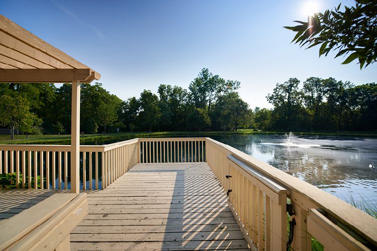 A wooden deck overlooking a serene lake with a fountain and surrounding trees in bright sunlight.