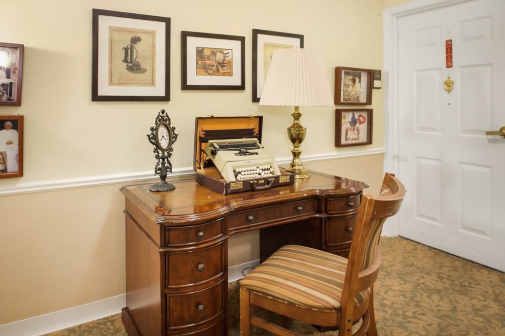 A vintage office setup with a typewriter on a wooden desk, accompanied by framed art and a classic lamp.
