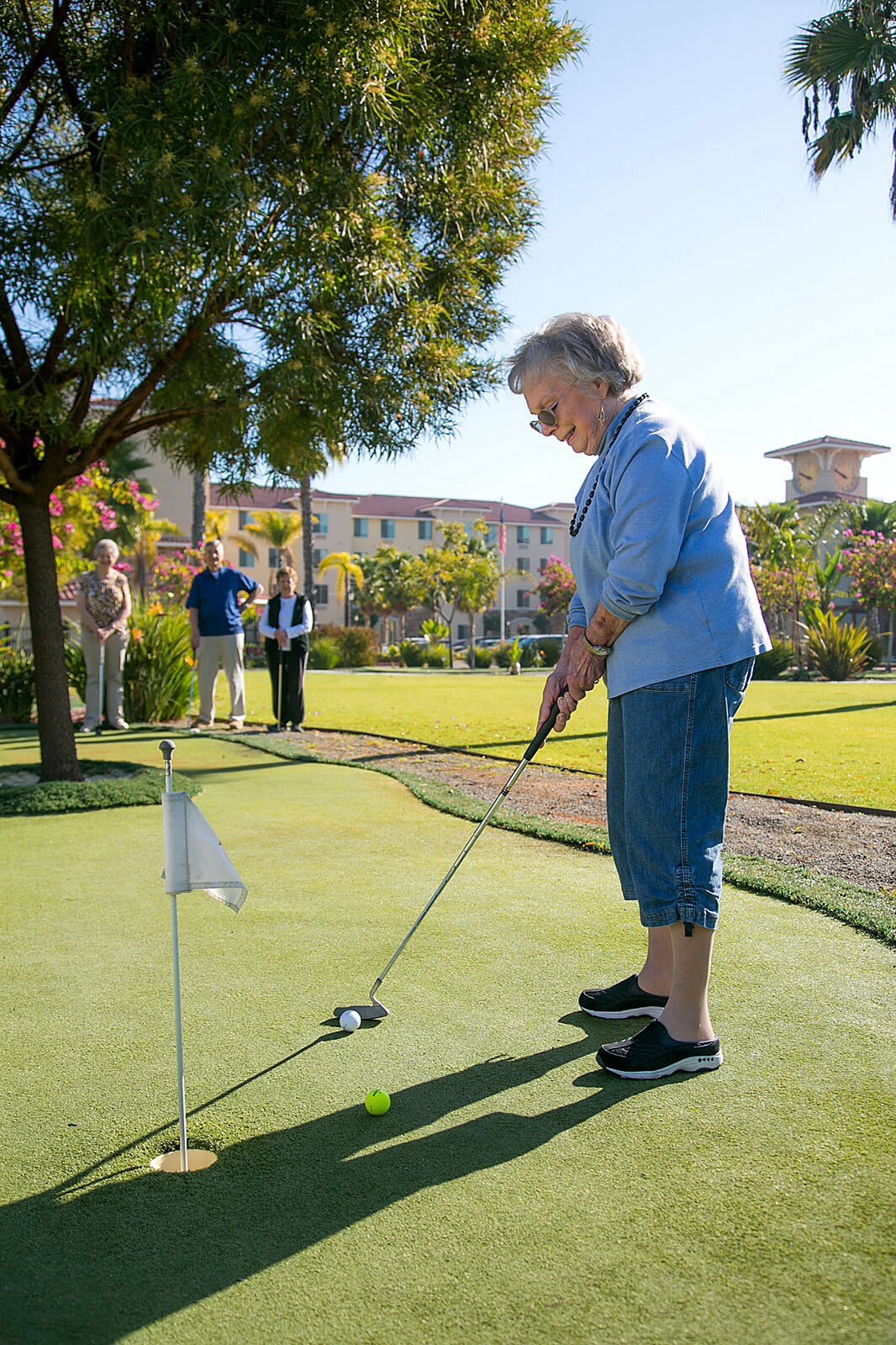 An elderly woman enjoys a sunny day playing golf, focusing on putting on a green course.