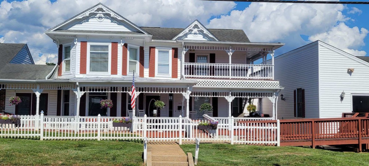 A two-story house with a porch and balcony, featuring a white picket fence and an American flag under a cloudy sky.