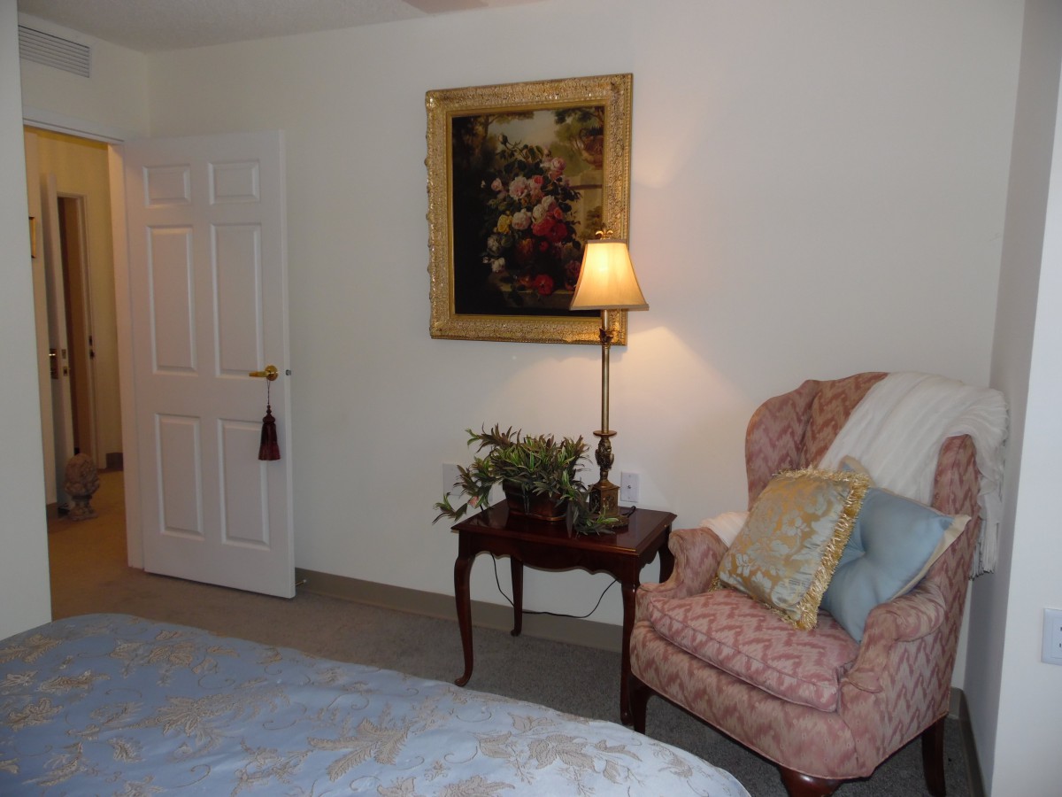 A cozy bedroom corner with an armchair, side table, and lamp under a framed painting.