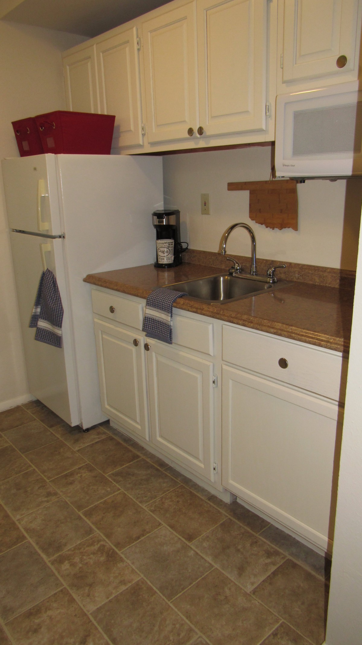 A small kitchen area featuring a refrigerator, sink, countertop, and cabinets with a coffee maker and towels.
