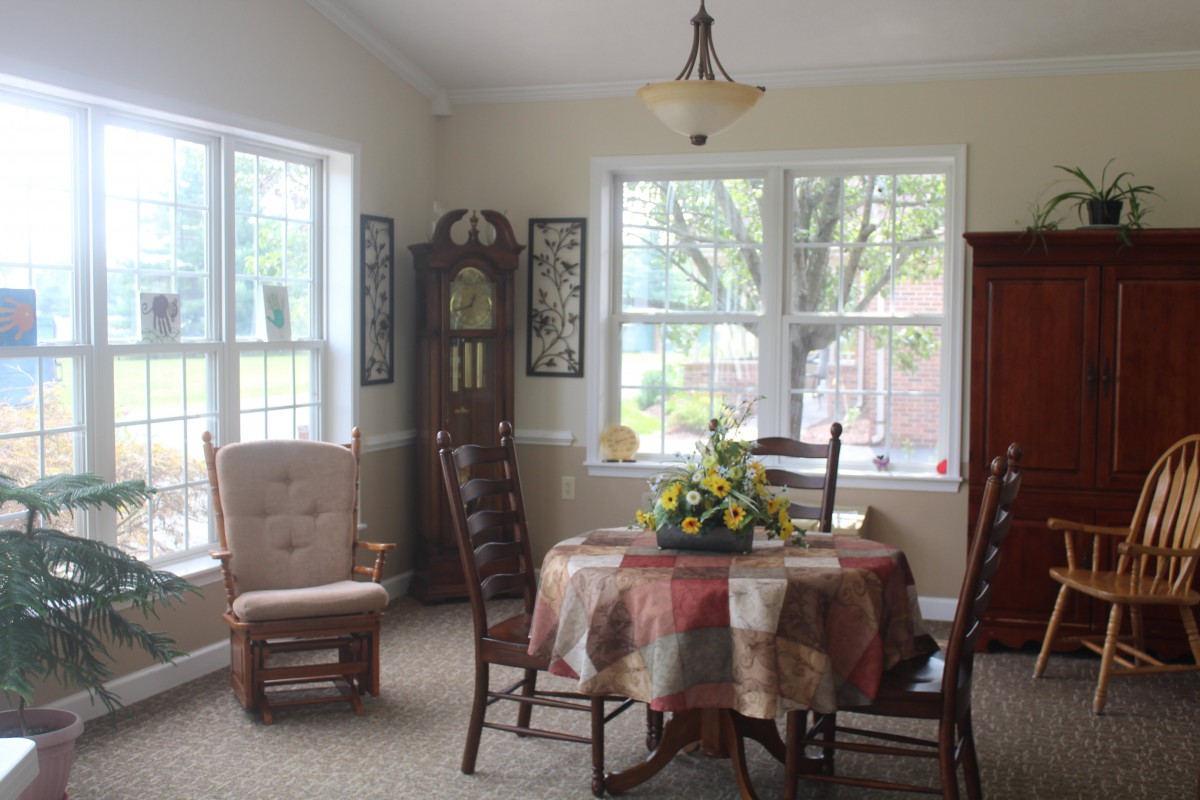 A cozy dining room with a table, chairs, and a grandfather clock, illuminated by natural light from large windows.