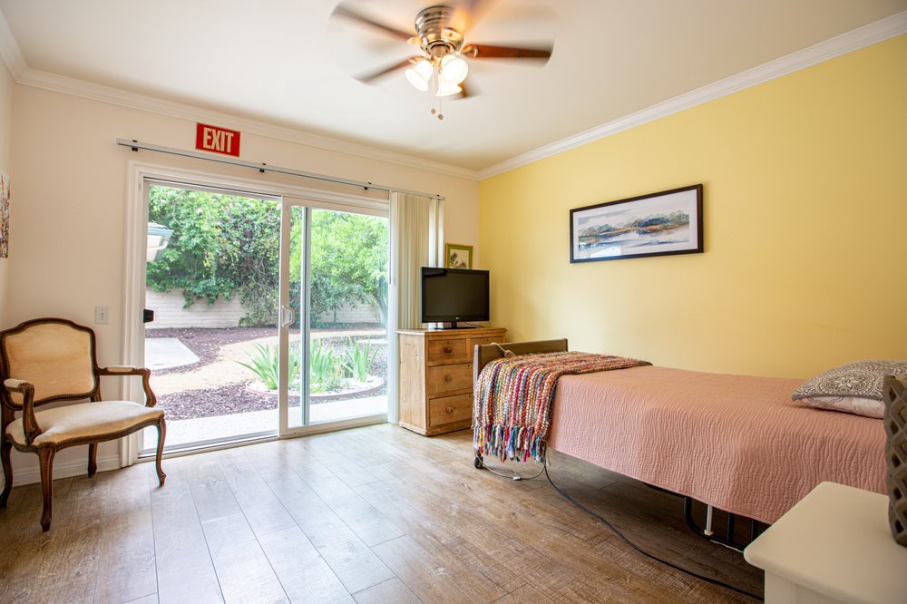 A modern and minimalist bedroom interior with ample natural light, featuring a single bed, wooden flooring, and a view of the garden through sliding glass doors.
