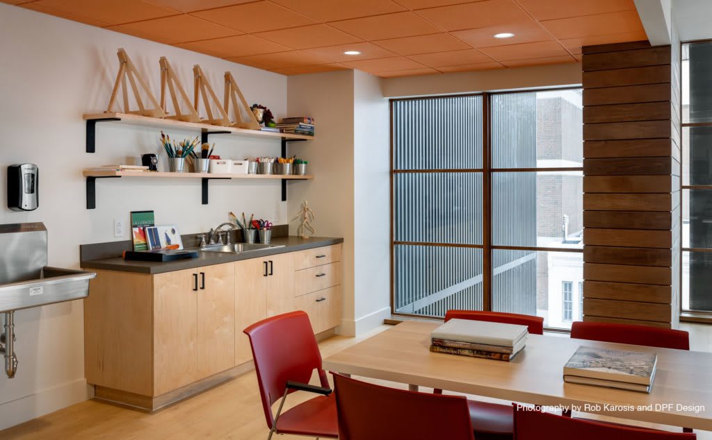 A modern office workspace featuring a table with red chairs, shelves with decorative items, and large windows providing natural light.