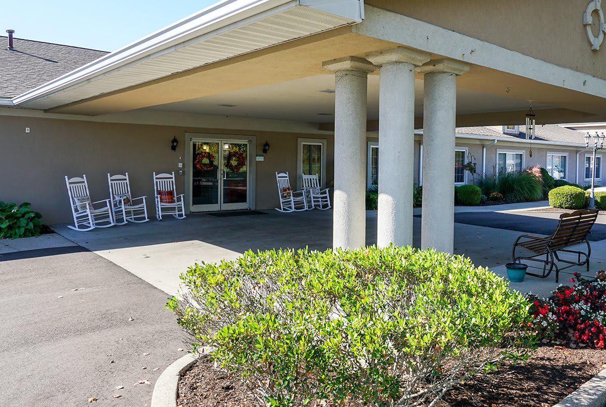 The image shows the entrance of a building with a covered porch, columns, and rocking chairs. A shrub is visible in the foreground.