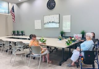 A spacious meeting room with a few seniors seated around tables, decorated with flowers and artwork, featuring a large clock on the wall.