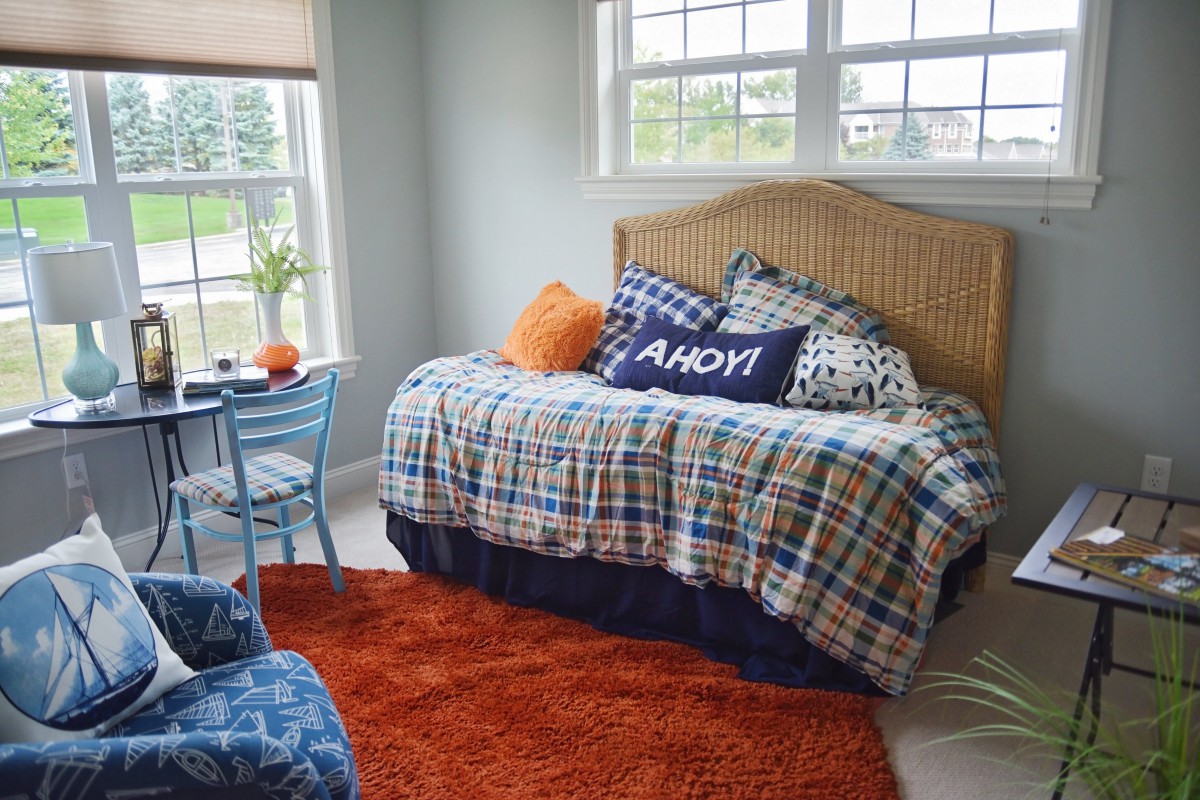A cozy bedroom featuring a plaid-bedecked daybed with nautical-themed pillows, an orange rug, and a small table next to a chair by the window.