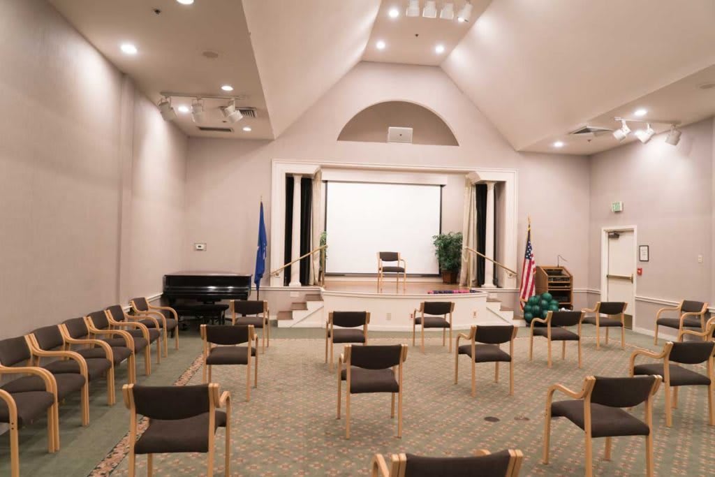 An auditorium with a stage set up for a presentation, featuring empty chairs, a projector screen, flags, and a piano.