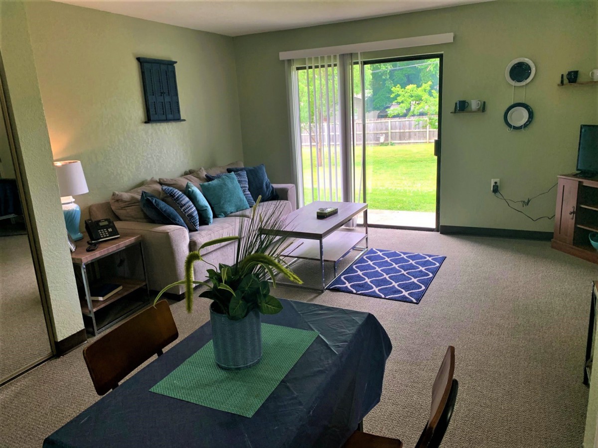 A cozy and well-lit living room with a sofa, coffee table, and dining area, opening to a green outdoor space through sliding glass doors.