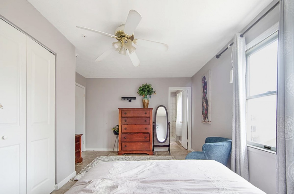 A cozy bedroom featuring a wooden dresser, an oval floor mirror, and a comfy blue chair.