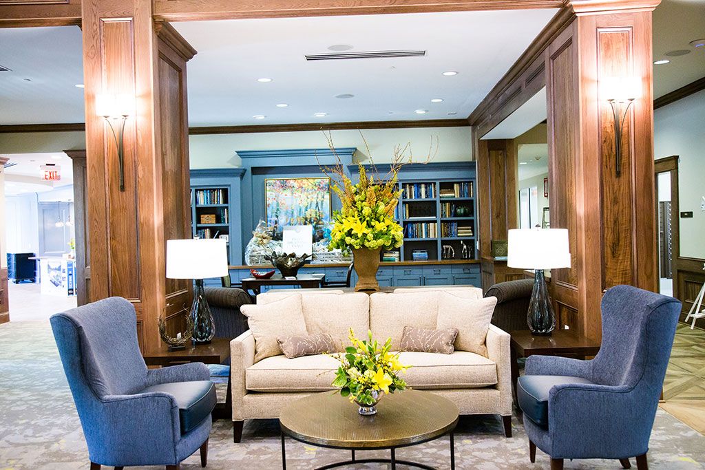 A cozy living room with a beige sofa, two blue chairs, and a coffee table adorned with floral arrangements, set against a backdrop of stylish bookshelves.