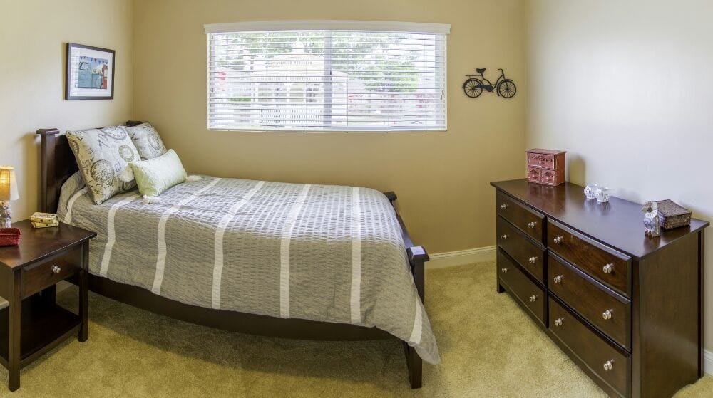 A cozy bedroom featuring a single bed with striped bedding, a window with blinds, and a wooden dresser.