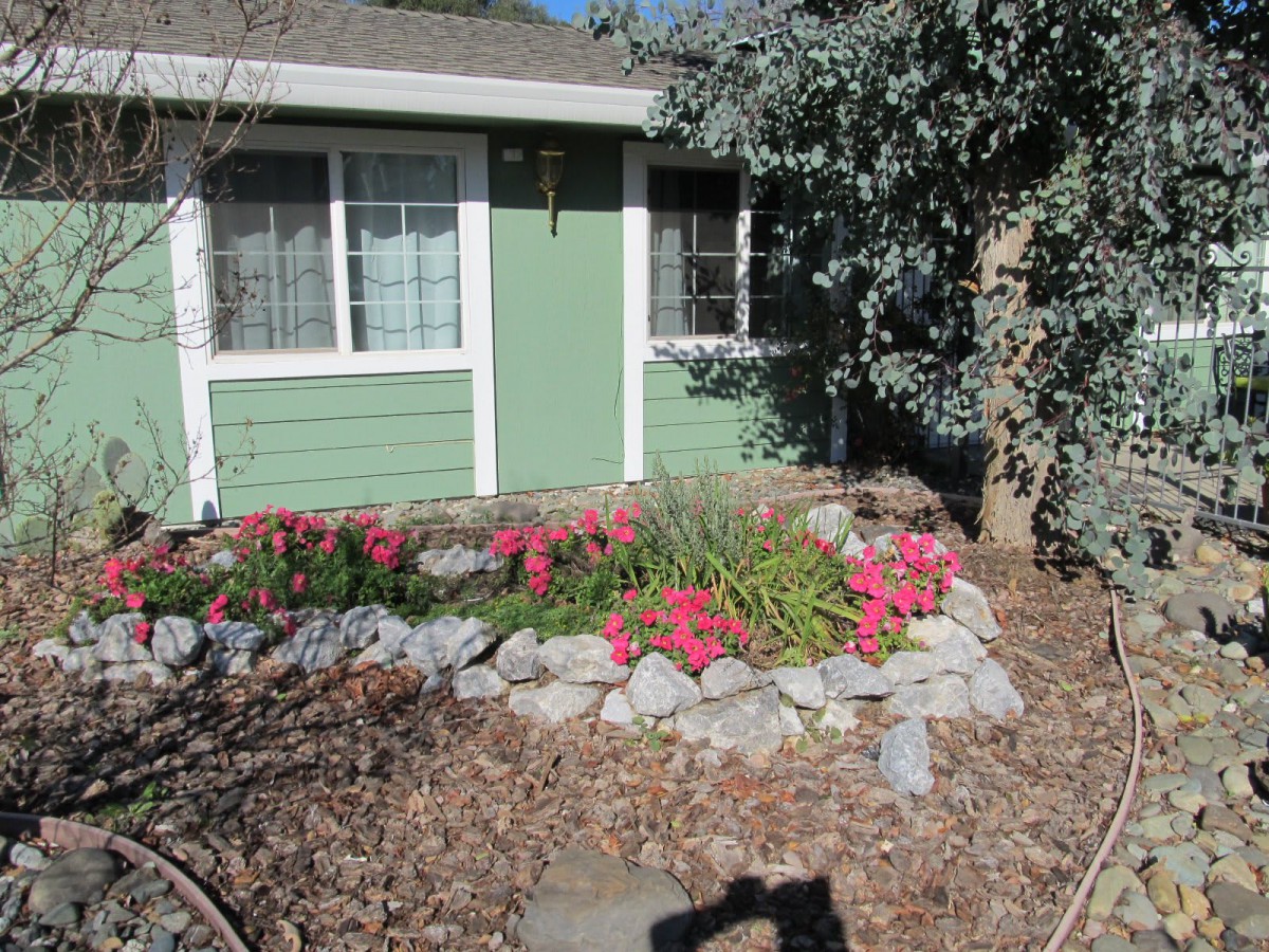 A charming garden with pink flowers and a rock border in front of a green house.
