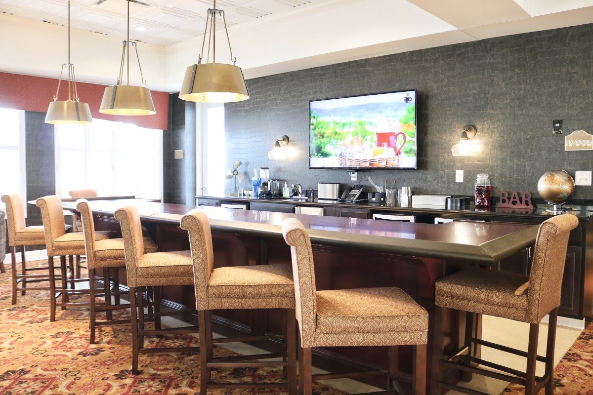 A cozy bar area featuring a long counter with high chairs, pendant lighting, and a large TV on the wall.