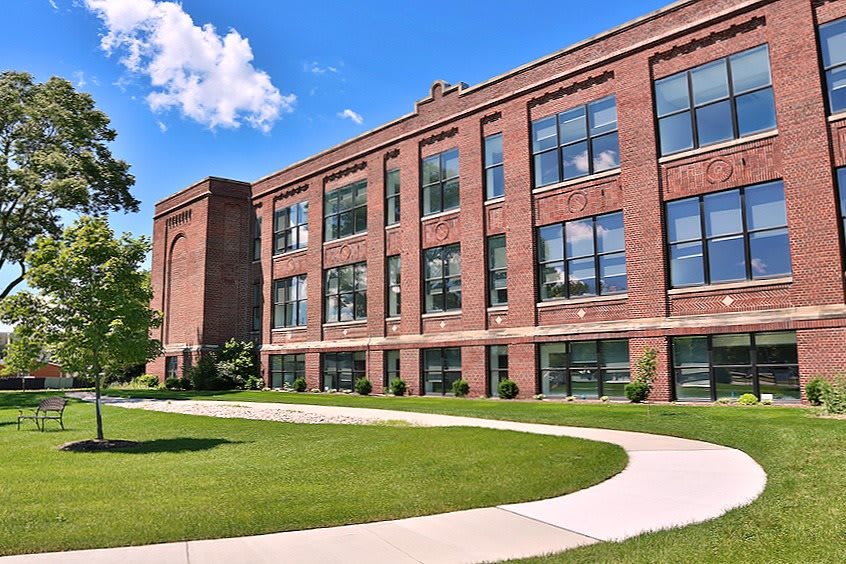 A large brick school building with numerous windows, surrounded by a green lawn and clear sky.