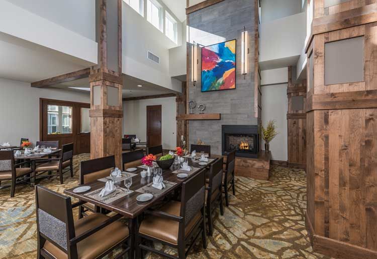 A modern dining room features a long table with elegant place settings, highlighted by large windows and an abstract painting above the fireplace.