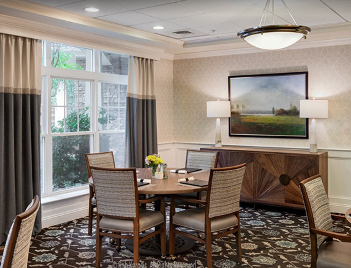 A cozy dining room with a table set for four, featuring elegant decor and ample natural light from large windows.