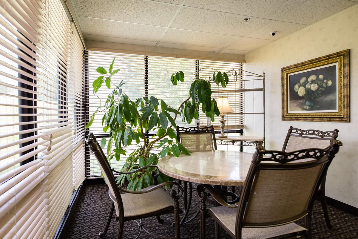 A cozy dining room with a round table, surrounded by chairs and lush indoor plants near large windows.