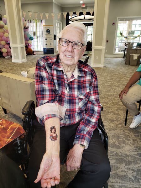 An elderly person with glasses shows off a new tattoo on their arm while sitting in a wheelchair indoors.