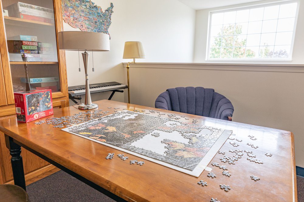 A cozy room with a puzzle in progress on a wooden table, accompanied by a lamp, chair, and a map on the wall.