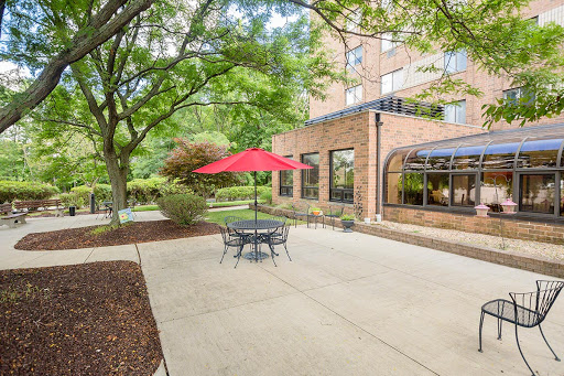 A serene outdoor patio area featuring a round table with chairs, shaded by a bright red umbrella, surrounded by lush greenery and a brick building.