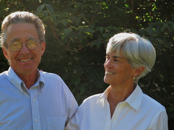 An elderly couple smiling at each other in a sunny outdoor setting.