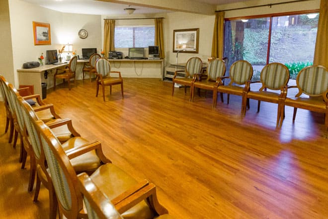 A spacious waiting room with wooden chairs lined against the walls, featuring a hardwood floor and large windows with curtains.
