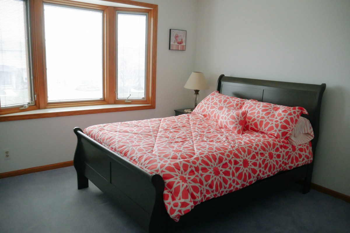 A cozy bedroom with a patterned red bedspread and natural light from a large window.