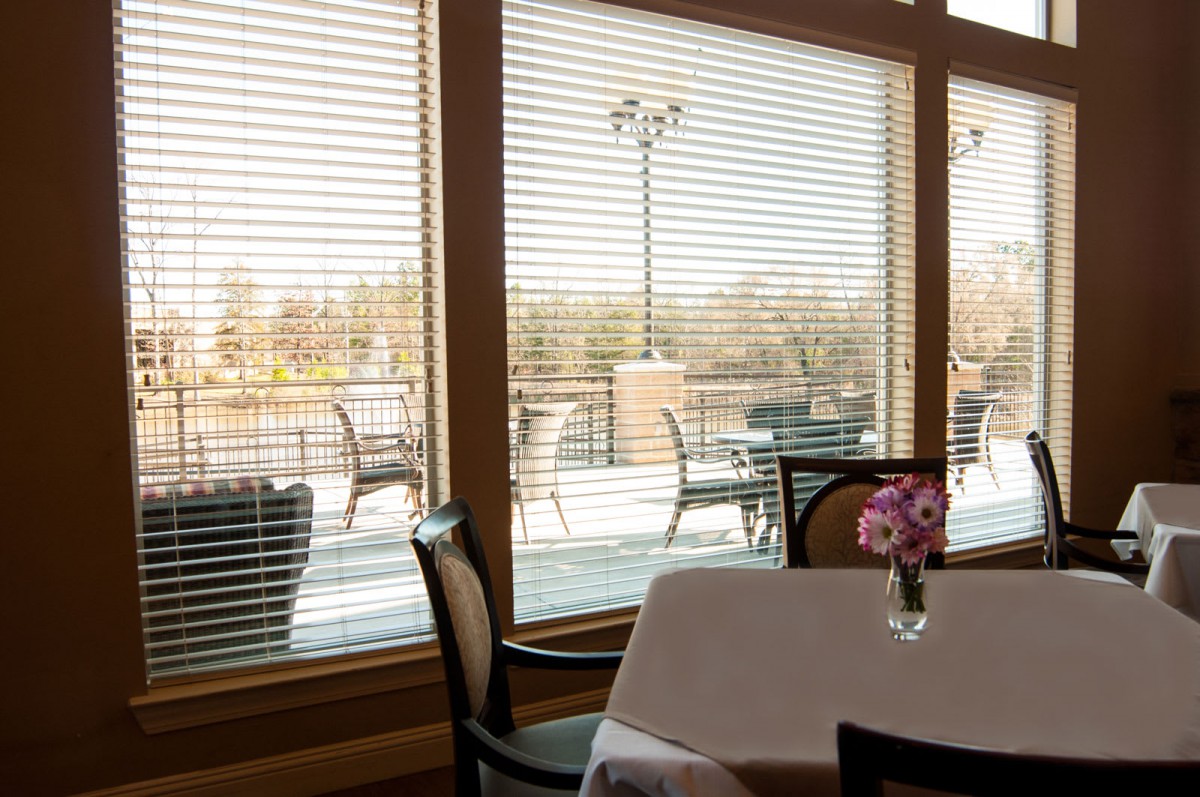 A cozy restaurant interior with a view of outdoor seating through large windows with blinds.