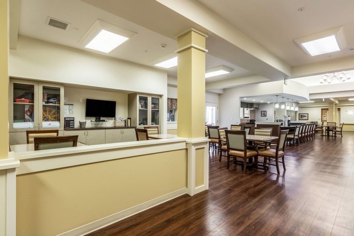 A spacious dining area with wooden floors, tables, and chairs, featuring a television and bright overhead lighting.