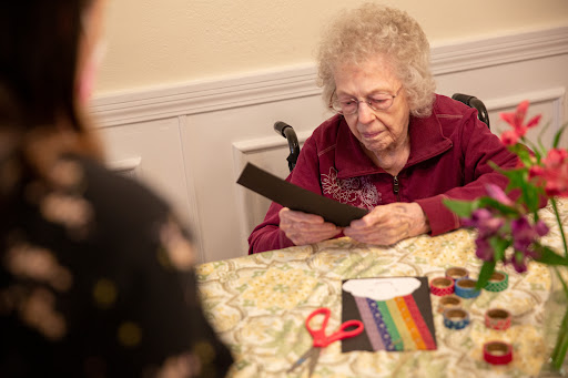 An elderly woman engaged in a crafting activity, focusing on colorful paper materials and scissors at a table.