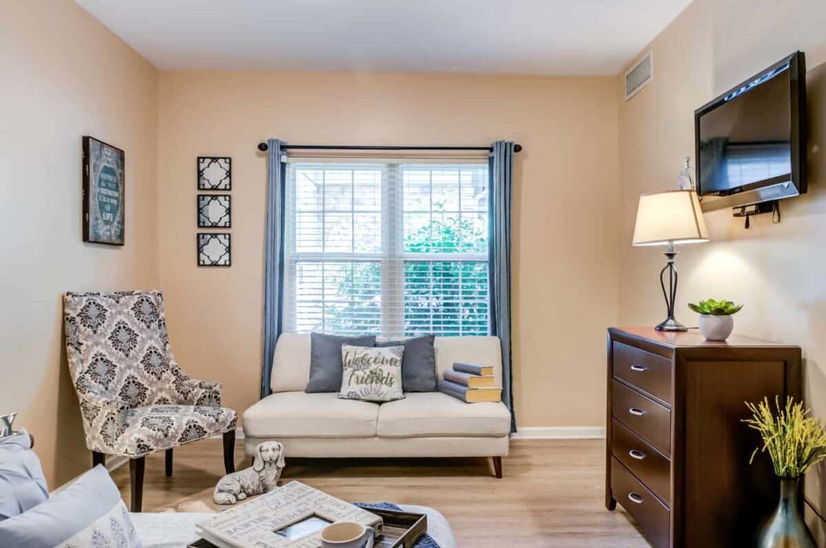 A cozy living room with a sofa, patterned armchair, and a tall lamp beside a window with light curtains.