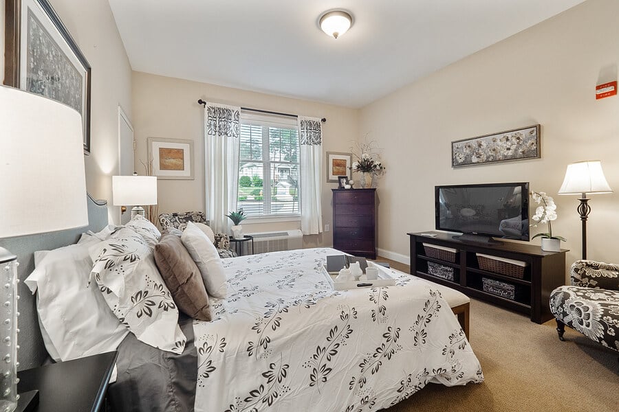 A cozy bedroom with a neatly made bed, TV stand, and large window letting in natural light.