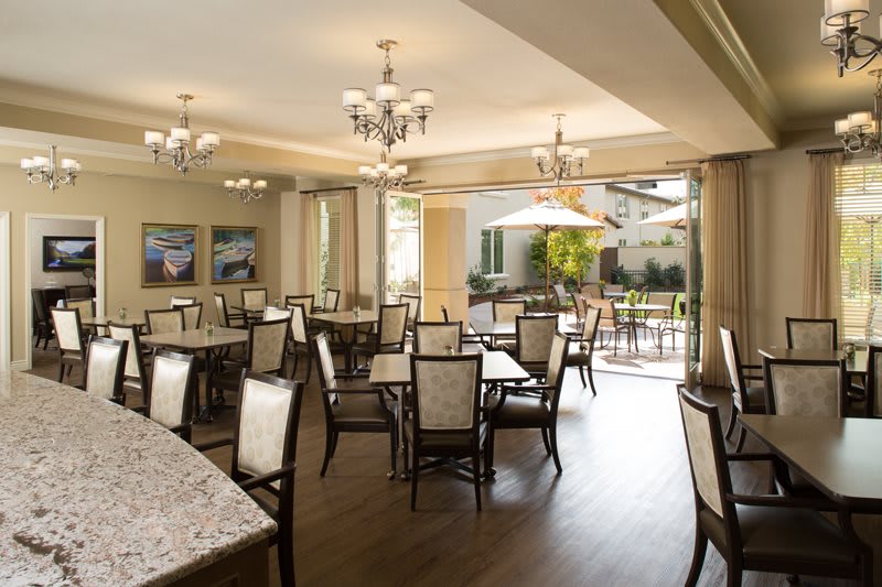 An elegant dining area featuring multiple tables and chairs, illuminated by chandeliers and open to a bright outdoor space.