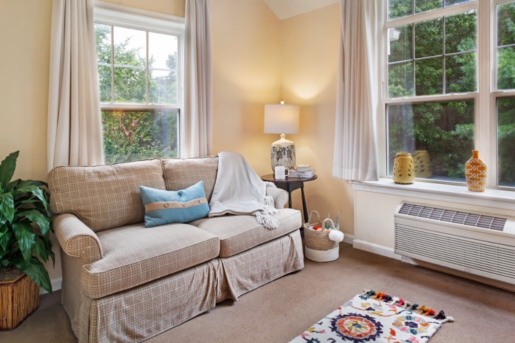 A cozy living room with a sofa, decorative cushions, a lamp on a side table, and natural light from large windows.