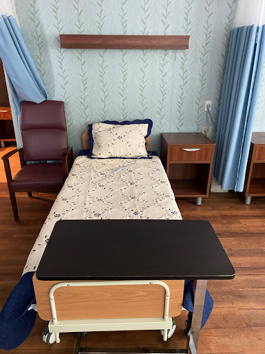 A clean and organized hospital patient room featuring a bed, bedside table, chair, and a writing surface.