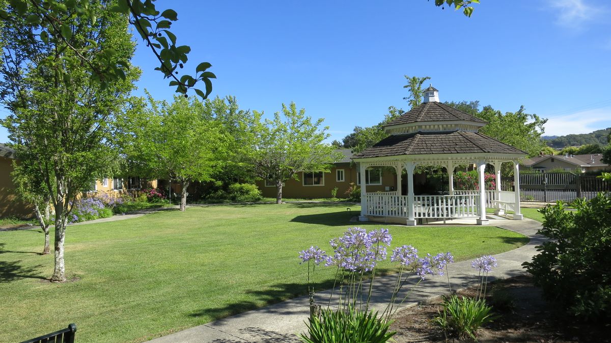 A peaceful garden scene with a white gazebo surrounded by lush greenery and blooming flowers under a clear blue sky.