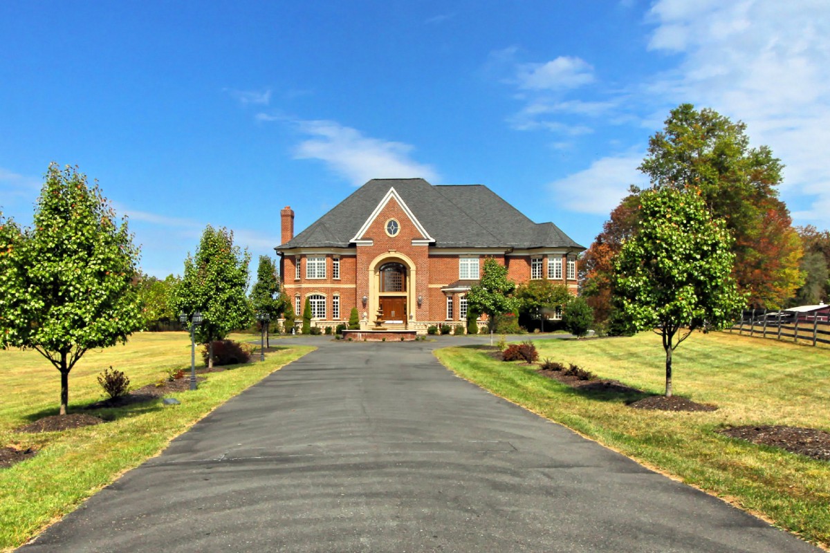 A large brick mansion at the end of a long driveway, surrounded by well-maintained landscaping and trees under a clear blue sky.