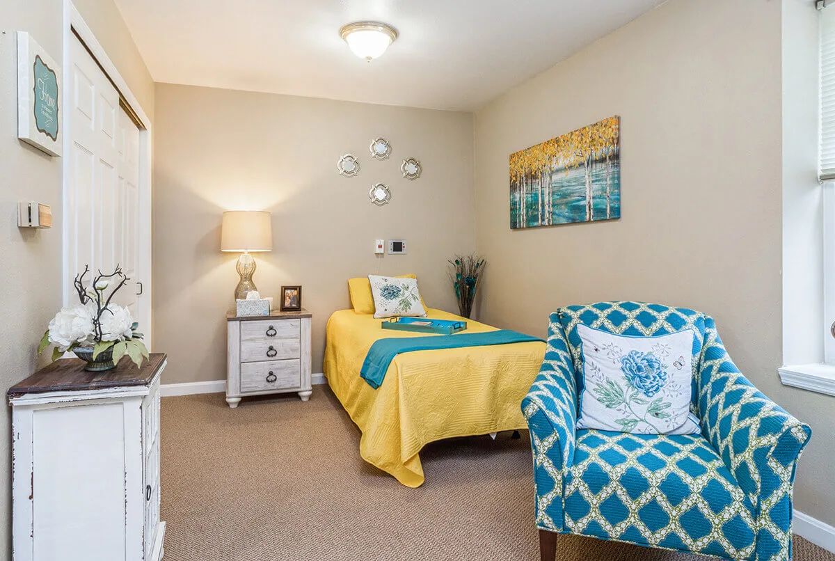 A cozy bedroom featuring a single bed with yellow and blue bedding, a patterned armchair, and decorative artwork on the wall.