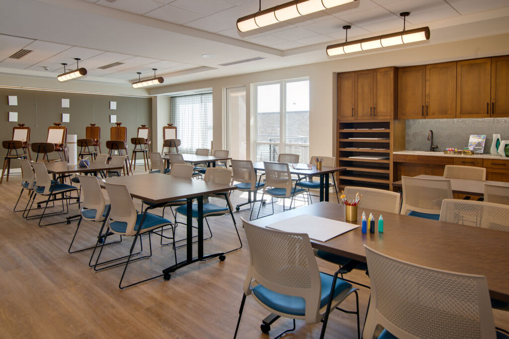 An art classroom with tables, chairs, and easels, illuminated by natural daylight.