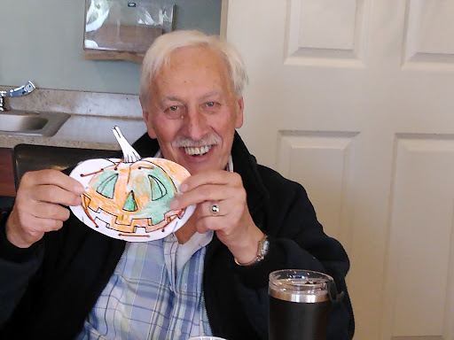 An elderly man proudly displaying his colorful pumpkin craft at a cozy indoor setting.