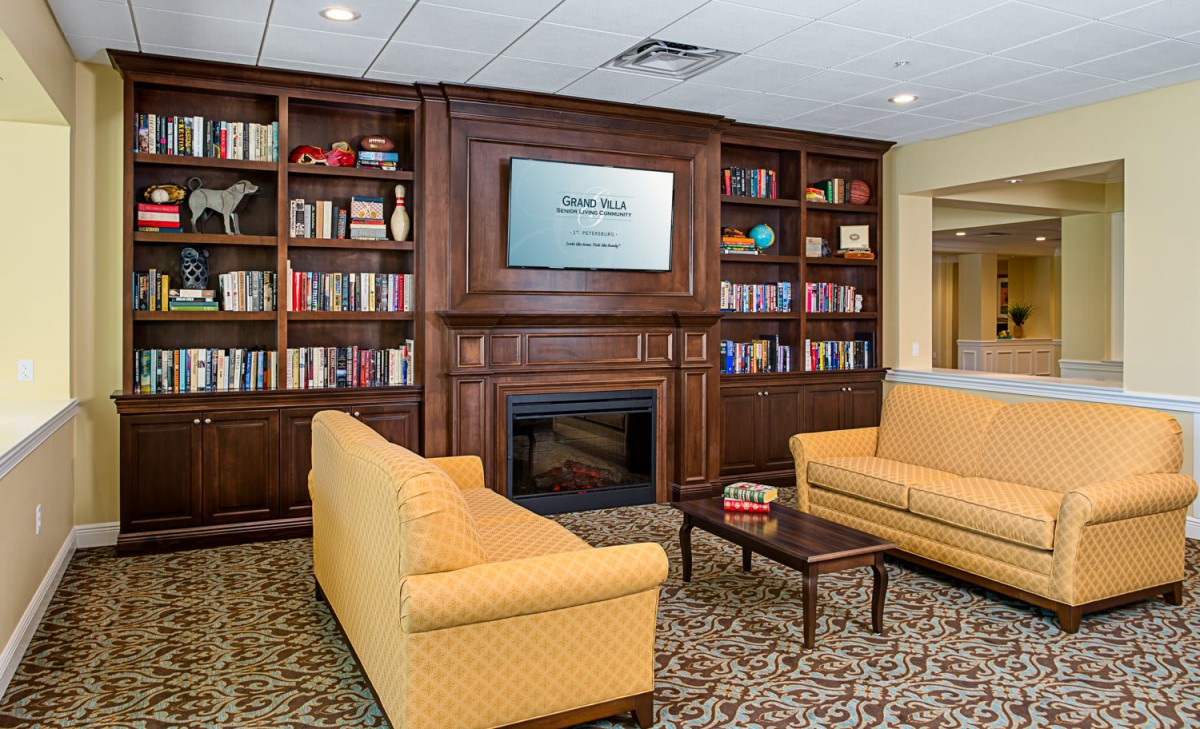 A cozy library room featuring wooden bookshelves, a television above the fireplace, and comfortable yellow couches.