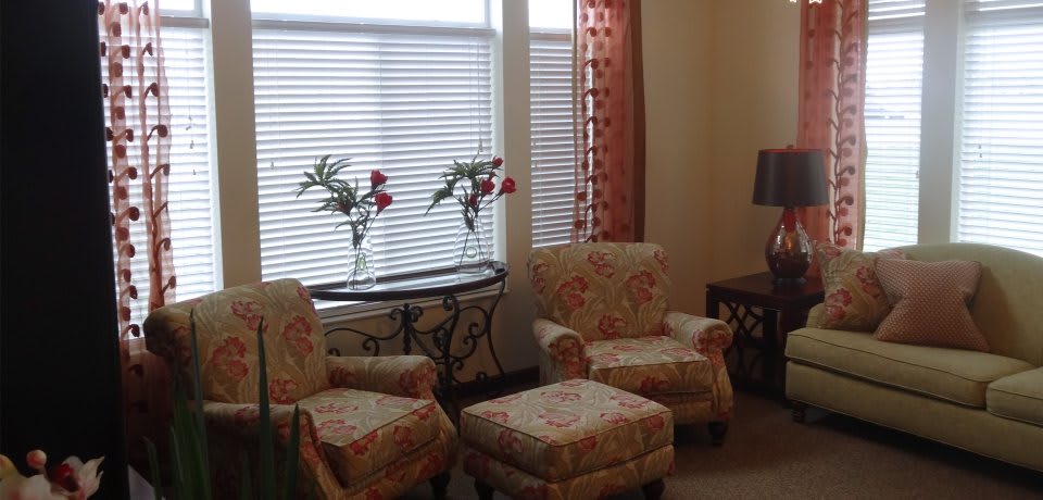 Cozy living room with floral-patterned chairs, matching ottomans, and a decorative table adorned with vases of flowers by the window.