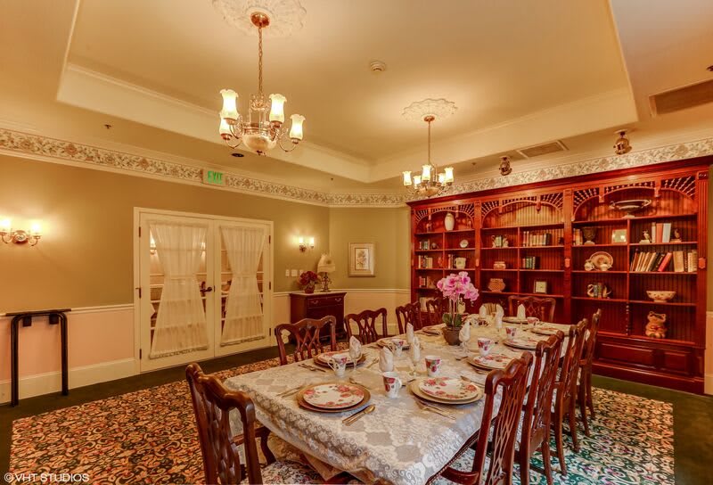 A beautifully decorated dining room with traditional wooden furniture, elegant table settings, chandeliers, and bookshelves lining the wall.
