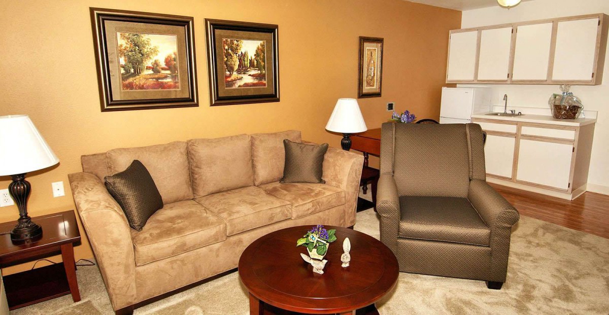 A cozy living room with a sofa, armchair, and coffee table, decorated with paintings and a lamp.