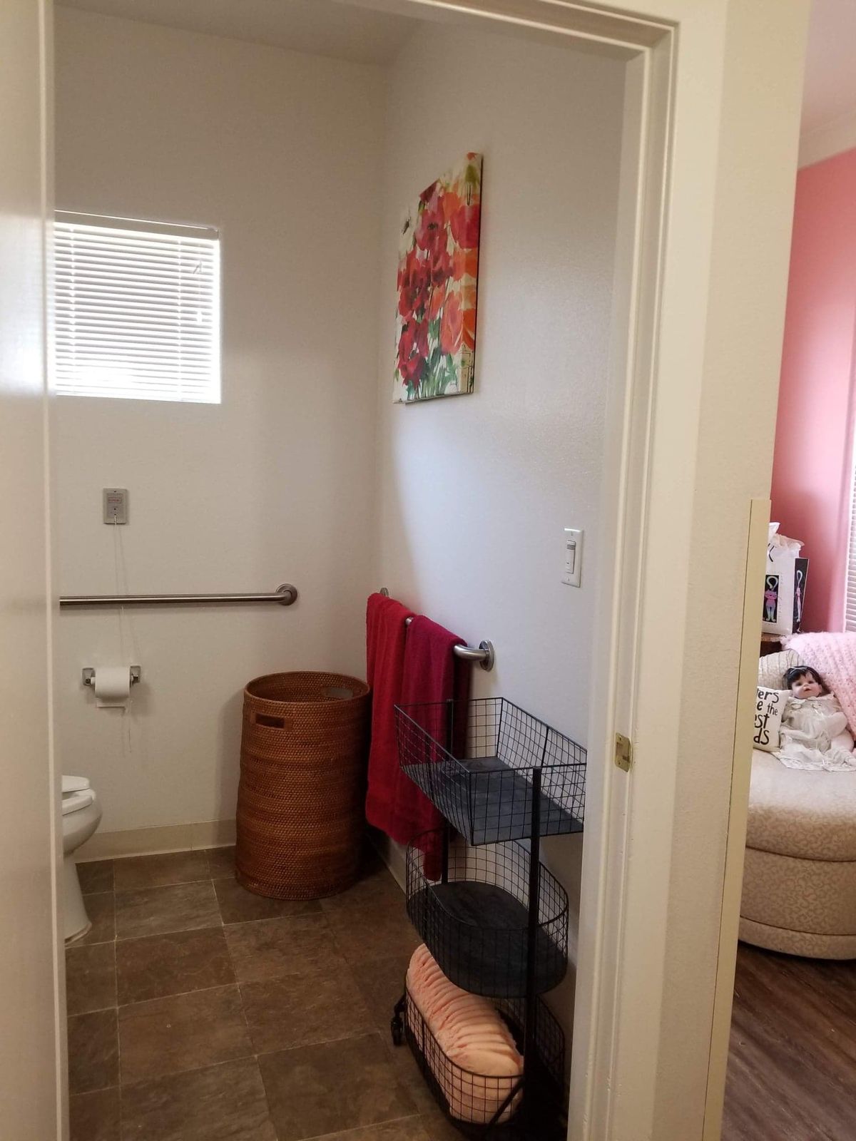 A view of a bathroom featuring a toilet, towel rack, and laundry basket, with a painting on the wall and a glimpse into an adjoining room.