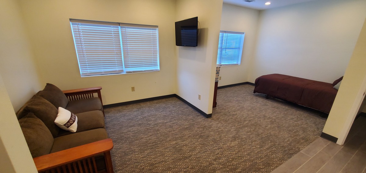 A spacious hotel room with a mounted television, multiple windows, and carpeted flooring.