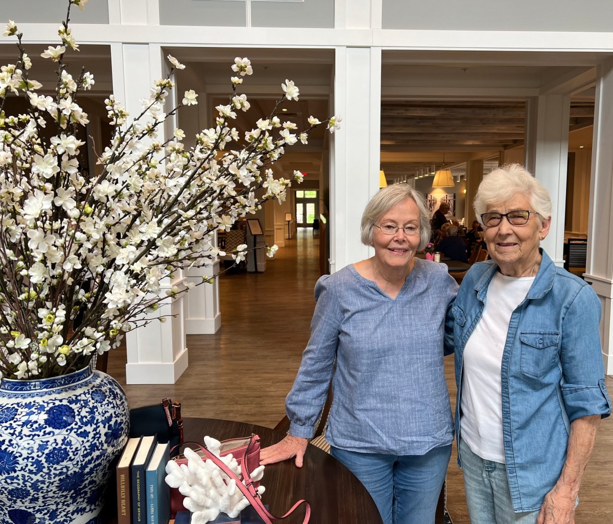 Two people smiling indoors next to a table with flowers and books.