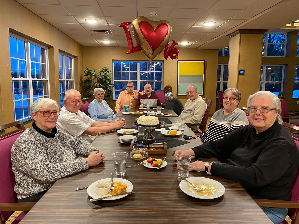 A group of friends gathered around a table for dinner, with heart-shaped balloons overhead.